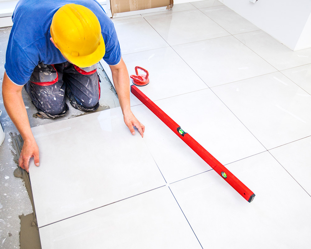 A man wearing a hard hat works diligently on laying tiles for a bathroom remodel, showcasing renovation efforts.