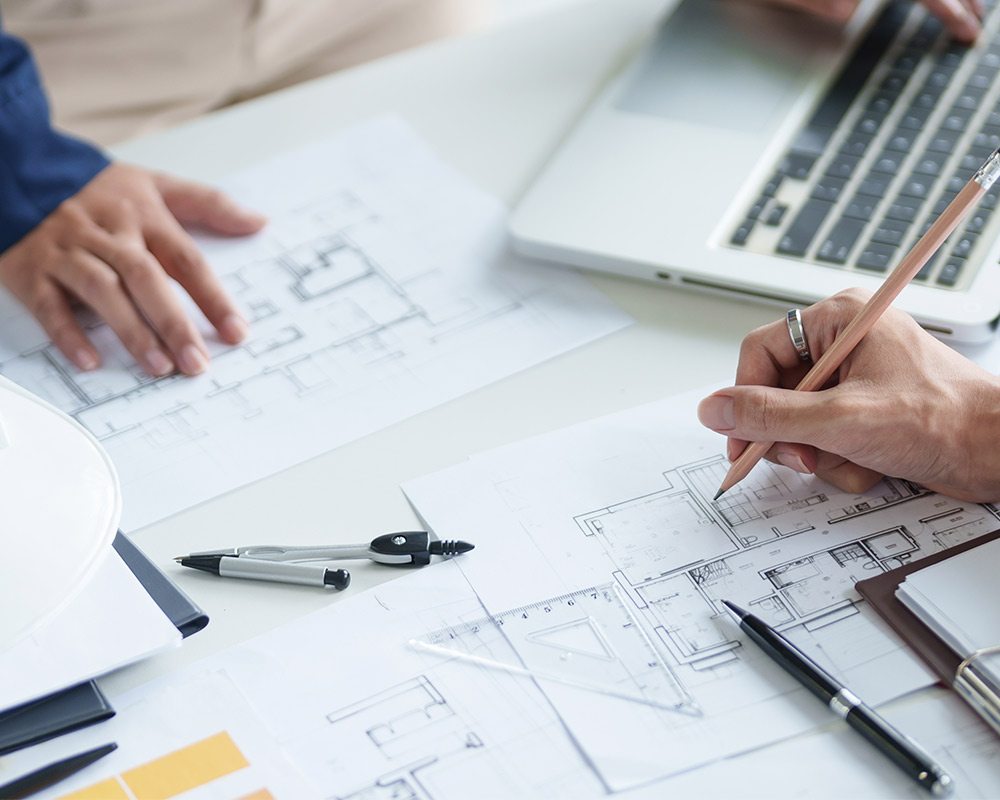 A man works on a laptop while sketching designs on paper, focused on custom home building in Barrington.
